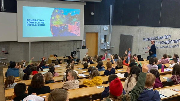 Ein Hörsaal voller Kinder, einige heben die Hände, der Dozent mit Mikrofon in der Hand läuft nach vorne zur Präsentation.