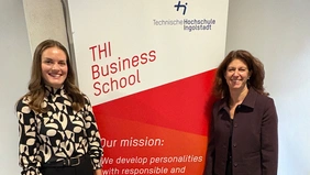 Zwei Frauen stehen vor einem Werbe-Roll-Up der THI Business School und lächeln in die Kamera.
