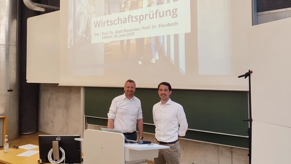 Zwei Männer lächeln in die Kamera, hinter Ihnen die Tafel und Leinwand des Hörsaals. 