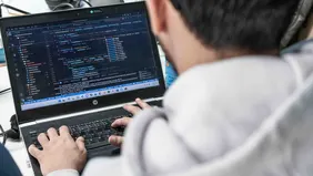 Man sieht über die Schulter eines Mannes auf seinen Laptop, wo er etwas programmiert.