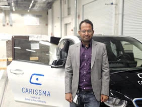 Für mehr Fahrzeugsicherhheit: Gerald Joy Sequeiras nutzt die CARISSMA-Halle für seine Fahrversuche (Foto: THI). Portrait mit Mann im Vordergrund, hinter Ihm ein Auto mit "CARISSMA"-Label auf der offenen Beifahrertüre