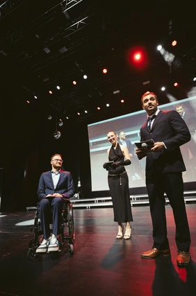Erst Paralympics-Bronze; dann den Abschluss: Lukas Gloßner (l.) mit dem Moderatoren-Duo (Foto: Simon Kahner). Student Lukas Gloßner mit zwei Moderierenden auf der Bühne bei der Akademischen Feier
