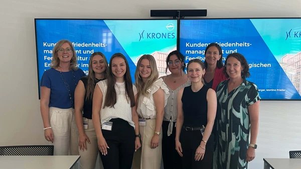 Eine Gruppe von acht Frauen lächelt in die Kamera, im Hintergrund zwei Fernseher mit der Startseite der Präsentation.