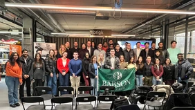: Ein Gruppenfoto im Veranstaltungsraum des brigk, in der Mitte der Gruppe wird die Flagge des Babson College gezeigt