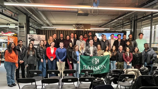 : Ein Gruppenfoto im Veranstaltungsraum des brigk, in der Mitte der Gruppe wird die Flagge des Babson College gezeigt