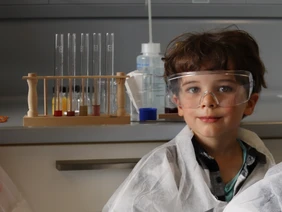Wie ein richtiger Wissenschaftler: Der 5-jährige Vincent im Labor (Foto: THI). KiGa-Kind in einem Labor mit Schutzbrille und -Kleidung, im Hintergrund Reagenzgläser