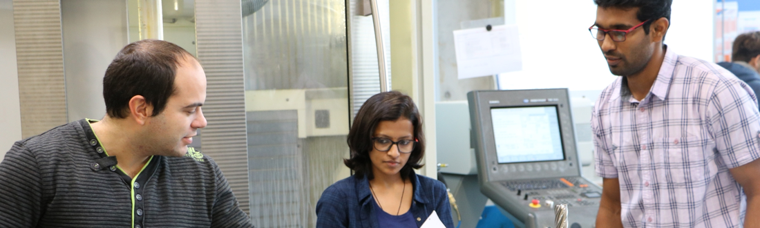 Drei Studenten stehen im Produktionsbereich beisammen, im Hintergrund ein Computer