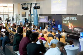 Volles Haus beim Gespräch mit Andreas Fraas (Foto: CoE). Bild vom Vortrag beim CAMP FIRE TALK mit Blick auf die Bühne und die Redner, die auf die Zuschauenden blicken